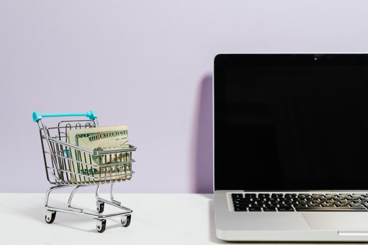 A small shopping cart holding a twenty-dollar bill sitting next to an open laptop, representing online holiday sales